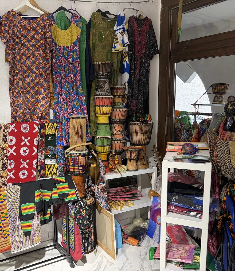 Bunte afrikanische Kleider und Stoffe, davor mehrere Trommeln.